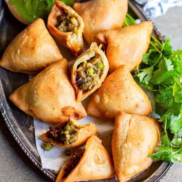 Golden-brown Indian samosas, some broken open to reveal savory filling, on a silver platter with cilantro.