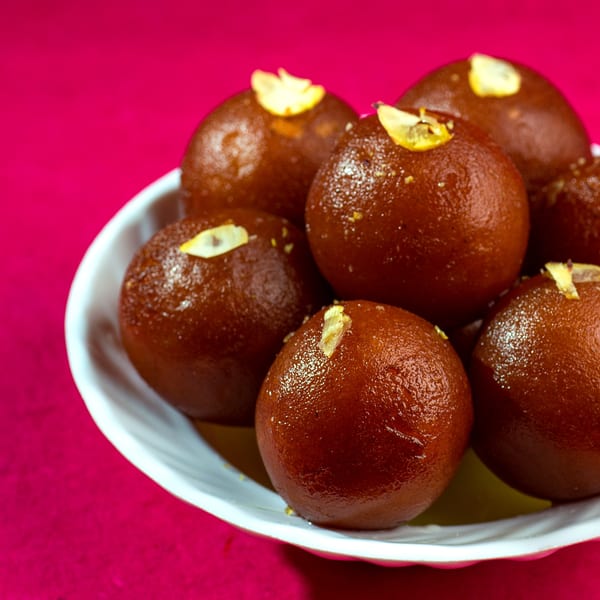 A white bowl filled with several glistening brown gulab jamun, garnished with slivered almonds.