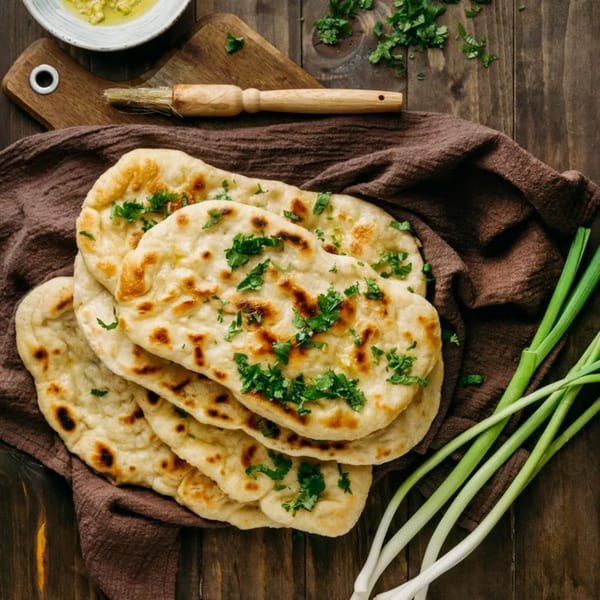 Golden-brown naan bread, garnished with fresh cilantro, on a brown cloth with green onions and a brush.